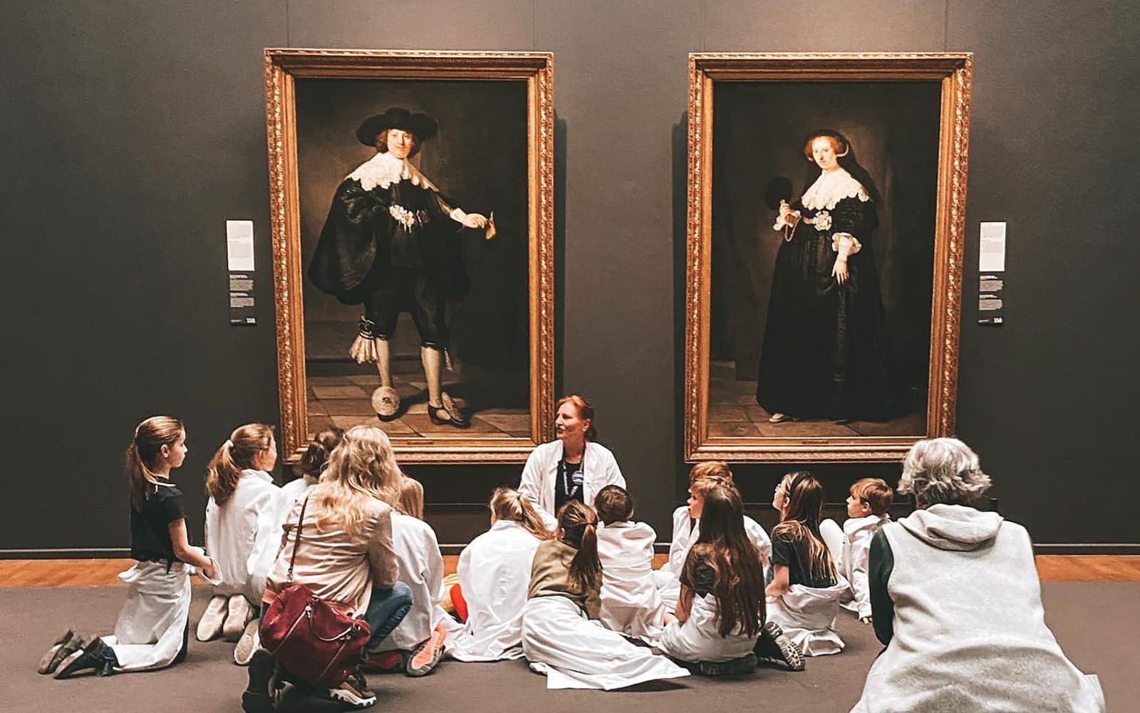 Mediadora a orientar um grupo de crianças sentadas frente a pinturas num museu